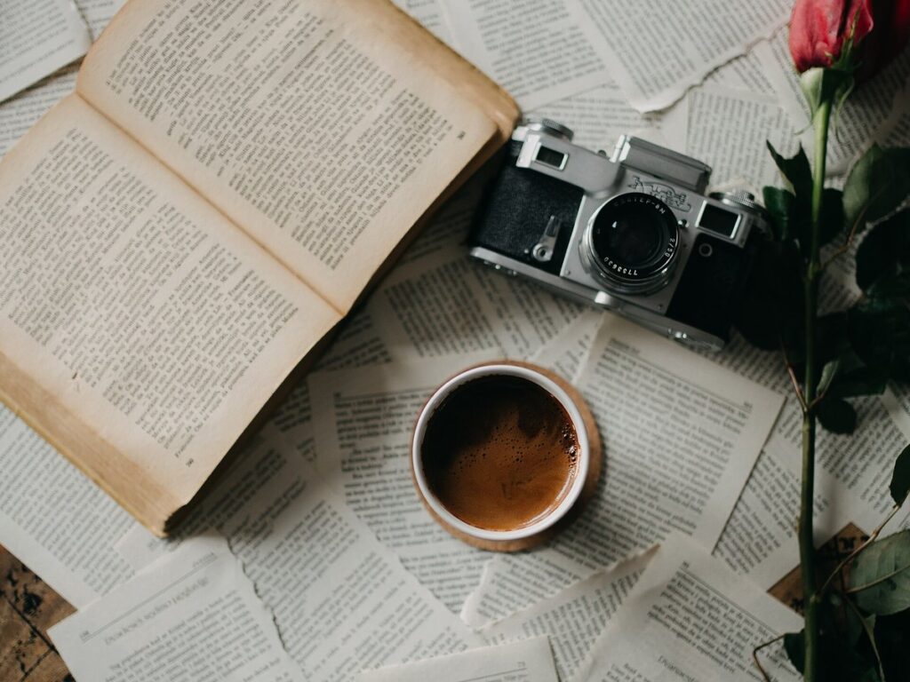 A warm and inviting setup features an open book surrounded by scattered pages, a vintage camera, a cup of coff